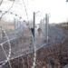Razor wire in the foreground frames a view of the border fence between Hungary and Serbia, with two guards waling along it