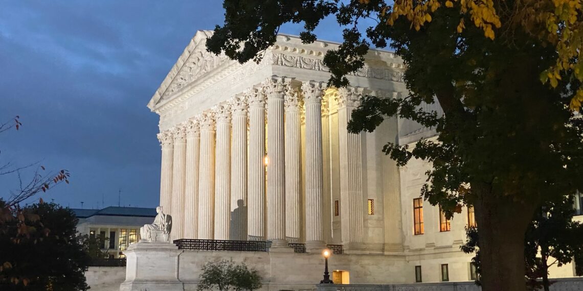 side view of supreme court building, illuminated against dark sky at dusk, with large tree in foreground