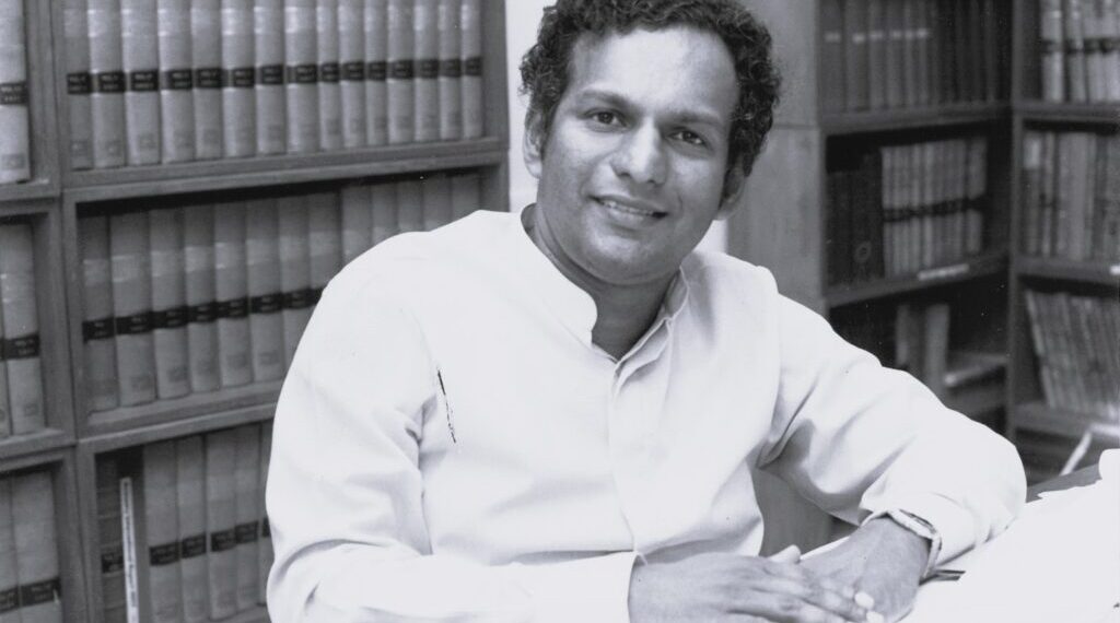 Black and white photo of Neelan Tiruchelvam sitting at a desk in front of book shelves smiling at the camera.