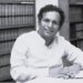 Black and white photo of Neelan Tiruchelvam sitting at a desk in front of book shelves smiling at the camera.