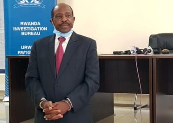 Paul Rusesabagina in a suit and tie, handcuffed, wearing a face mask pulled down around his chin, in front of a sign saying ‘Rwanda Investigation Bureau’