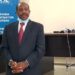 Paul Rusesabagina in a suit and tie, handcuffed, wearing a face mask pulled down around his chin, in front of a sign saying ‘Rwanda Investigation Bureau’