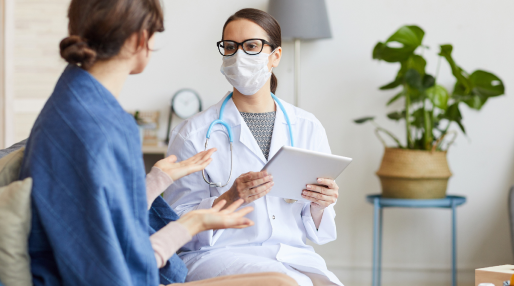 A doctor in a white coat and a mask holding a patient chart sitting across from a patient.