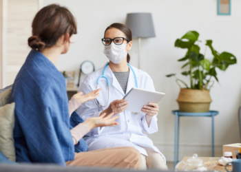 A doctor in a white coat and a mask holding a patient chart sitting across from a patient.