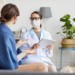 A doctor in a white coat and a mask holding a patient chart sitting across from a patient.