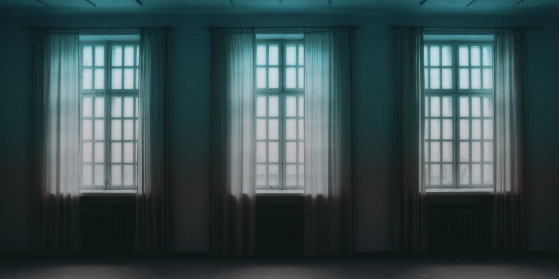 An empty room with three windows, all with long, sheer curtains. The two ceiling lights are off. Nothing but light can be seen outside the windows.