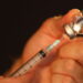 Two hands are using a syringe and needle to pull medication from a small glass vial.
