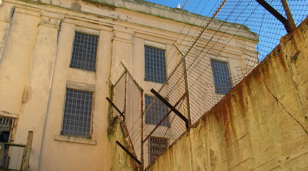 Photo of a beige building with high walls, at the top of the walls are fences with barbed wire.