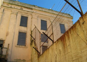 Photo of a beige building with high walls, at the top of the walls are fences with barbed wire.