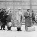 A black and white photo of men wearing clothing from the early 1900s. The men are carrying suit cases and standing in a line at Ellis Island after arriving in the US.
