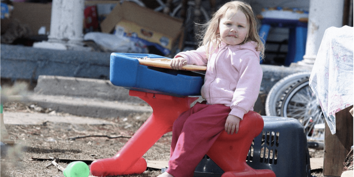 A child with no shoes, pink pants, and a light pink jacket sits on a red, blue, and yellow hard plastic toy. She is in a backyard surrounded by gray dirt, trash, and other junk. Her face is dirty, and her hair is messy.