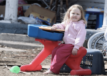 A child with no shoes, pink pants, and a light pink jacket sits on a red, blue, and yellow hard plastic toy. She is in a backyard surrounded by gray dirt, trash, and other junk. Her face is dirty, and her hair is messy.