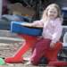 A child with no shoes, pink pants, and a light pink jacket sits on a red, blue, and yellow hard plastic toy. She is in a backyard surrounded by gray dirt, trash, and other junk. Her face is dirty, and her hair is messy.