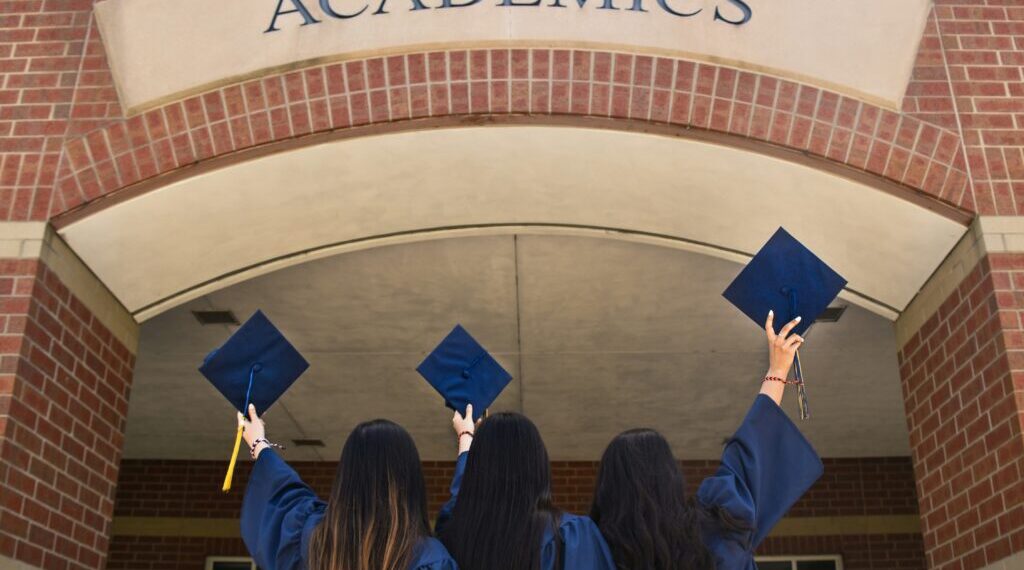 Three girls in blue graduation robes facing an Academics building holding up their caps.