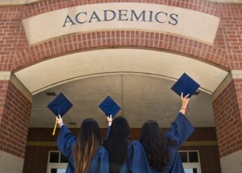 Three girls in blue graduation robes facing an Academics building holding up their caps.