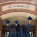 Three girls in blue graduation robes facing an Academics building holding up their caps.