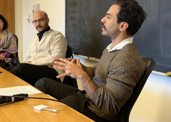 Speaker Nader Andrawos in conversation with moderator Omar Abdel-Ghaffar. They are both sitting at a table in front of a blackboard.