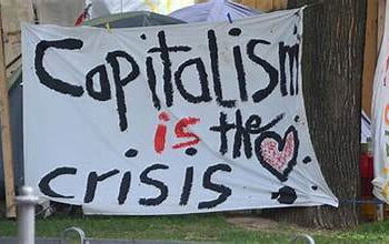 Painted sign that reads "Capitalism is the crisis" in black and red text.