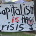 Painted sign that reads "Capitalism is the crisis" in black and red text.